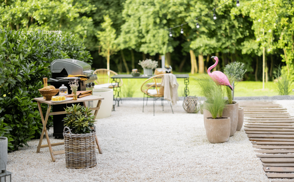 Barbecue ou plancha : espace extérieur avec gravier, chemin en bois à droite et jardin avec pelouse au fond. Barbecue avec table d'appoint, vases et plantes, salon de jardin, flamand rose